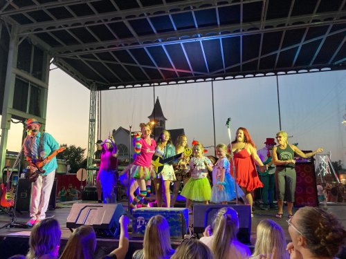 A lively stage performance with colorful performers, including clowns and children, during a sunset event.