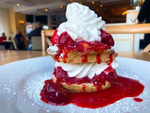 A stack of fluffy pancakes topped with whipped cream and strawberry sauce, dusted with powdered sugar on a white plate.