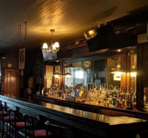 A dimly lit bar with a long counter, shelves of liquor bottles, and a vintage chandelier overhead.