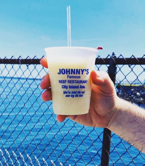A hand holding a cup from Johnny's Famous Reef Restaurant, with a waterfront view in the background.