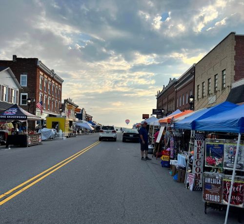 A street lined with vendor tents and parked cars, under a cloudy sky with a hot air balloon in the distance.