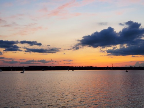 A serene sunset over a calm lake, with colorful clouds reflecting on the water and silhouettes of boats in the distance.