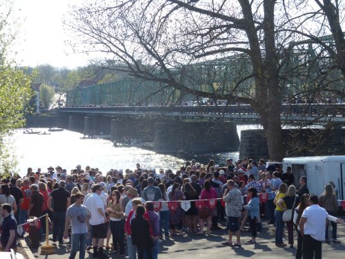 A large crowd gathers by a riverbank, with a bridge in the background and trees lining the area on a sunny day.