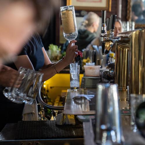 A bartender pours a drink into a glass filled with ice at a busy bar, with various bottles and tools in the background.