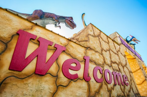A colorful "Welcome" sign with a dinosaur sculpture on top against a clear blue sky.