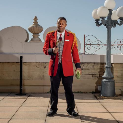 A man in a formal red jacket and black pants stands confidently outdoors, holding a microphone and smiling.