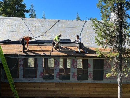 Three workers installing a roof on a house, surrounded by trees and clear blue sky.