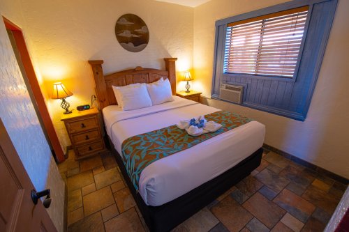 Cozy bedroom with a queen bed, wooden furniture, lamps, and a window with blinds. Decorated with tropical accents.