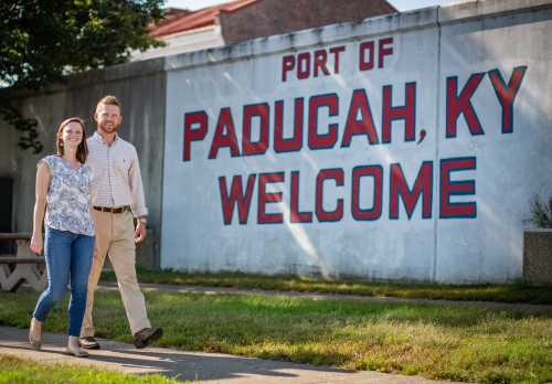 A couple walks together in front of a large "Port of Paducah, KY Welcome" mural on a sunny day.