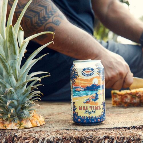 A can of Kona Brewing's Mai Time Light beer next to a fresh pineapple on a wooden surface.