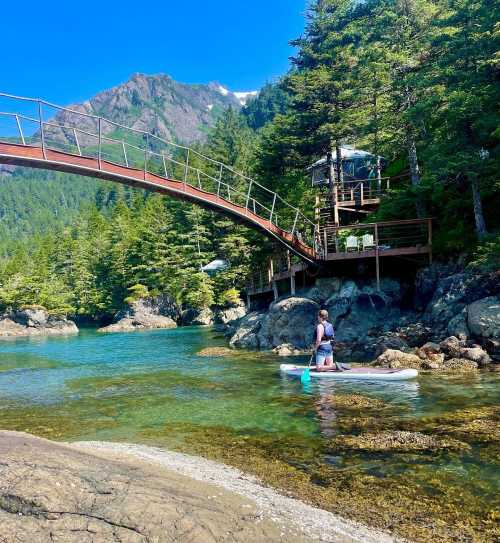 A person stands on a paddleboard in a clear blue lake, with a wooden bridge and lush green trees in the background.