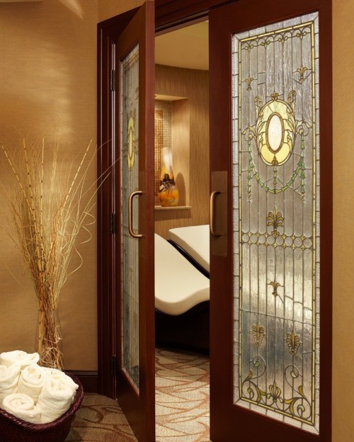 Elegant wooden doors with stained glass, leading to a serene spa area with rolled towels and soft lighting.