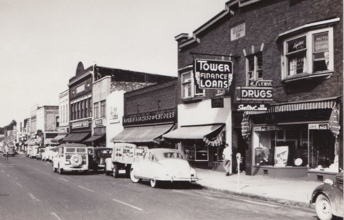 Black and white photo of a vintage street lined with shops, cars, and signs for finance and drug stores.