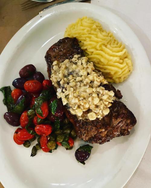 A plated meal featuring a steak topped with crushed nuts, served with mashed potatoes and a side of sautéed vegetables.