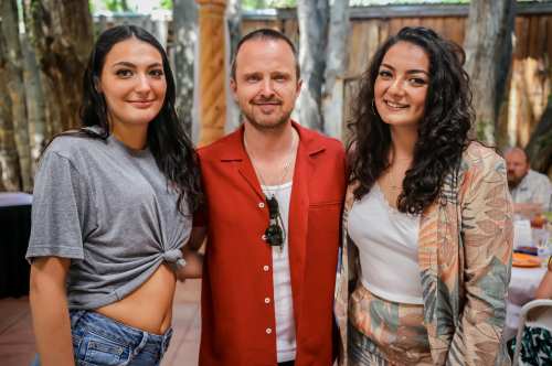 Three people pose together outdoors, smiling. Two women wear stylish outfits, and a man in a red shirt stands between them.