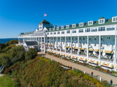 A grand white hotel with a green roof, overlooking a scenic landscape and blue sky, with people walking along the path.