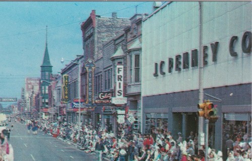 A busy street scene with a parade, featuring storefronts and crowds of people along the sidewalk.
