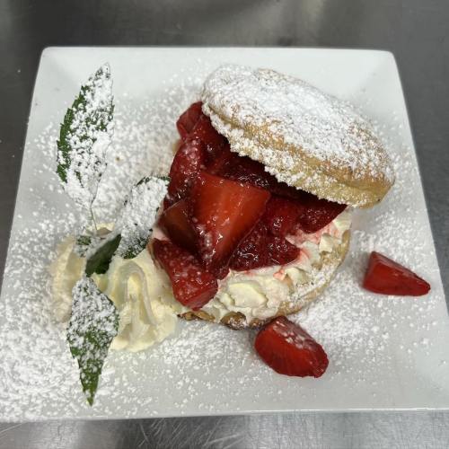 A dessert featuring a biscuit topped with whipped cream and fresh strawberries, dusted with powdered sugar and garnished with leaves.