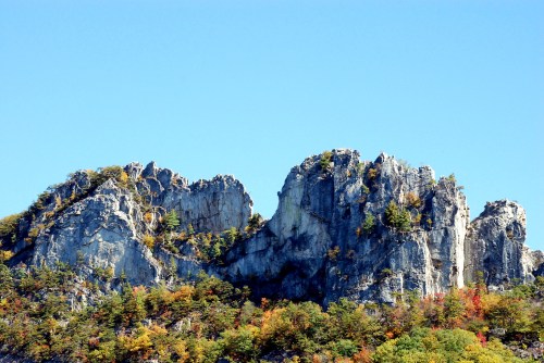 A rugged rock formation rises against a clear blue sky, surrounded by colorful autumn foliage.