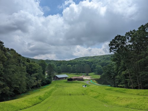 A lush green landscape with a pond, surrounded by trees and a rustic building under a cloudy sky.
