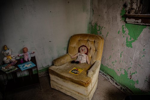 An old, worn yellow armchair in a dilapidated room, with a doll sitting on it and toys scattered nearby.