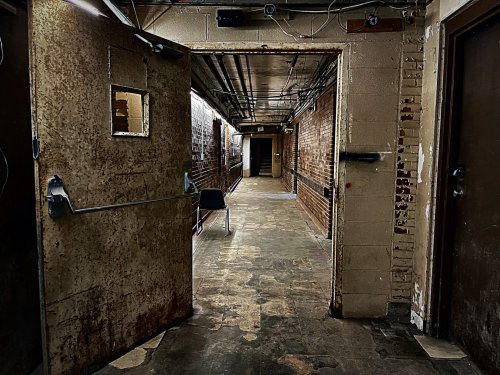 Dimly lit hallway with brick walls, a metal door ajar, and a single chair in the distance.