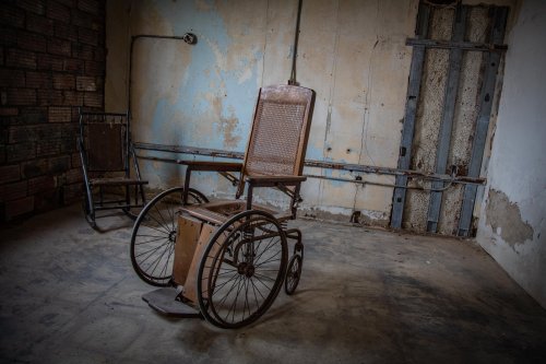 An old, wooden wheelchair sits in a dimly lit, abandoned room with peeling walls and a rusty door.