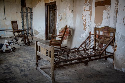 Abandoned room with a rusty bed frame and two old chairs, featuring peeling paint and a worn wooden floor.
