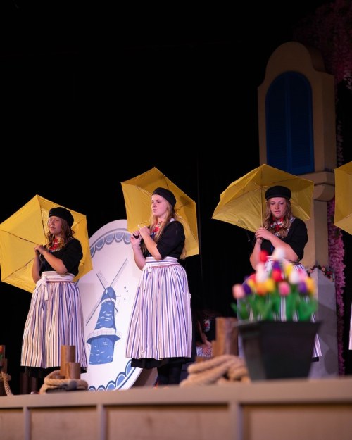 Three performers in black tops and striped skirts hold yellow umbrellas on stage, with a colorful flower pot in the foreground.