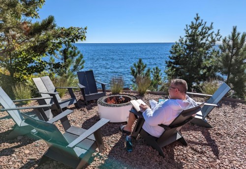 A person sits by a fire pit, reading a book with a view of the lake and trees on a sunny day.