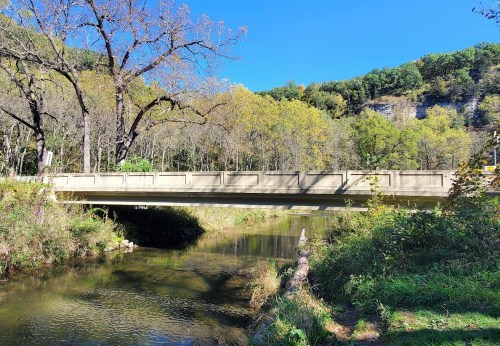 A concrete bridge spans a calm river, surrounded by trees and a hillside under a clear blue sky.