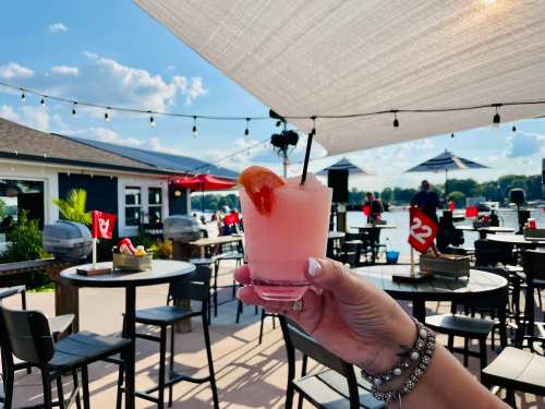A hand holds a pink cocktail with a cherry, set against a lakeside patio with tables and umbrellas.