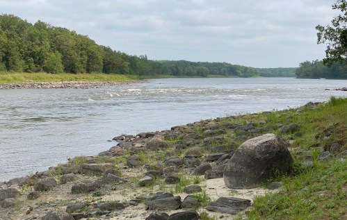 A calm river flows through a lush green landscape, with rocks lining the shore and trees in the background.