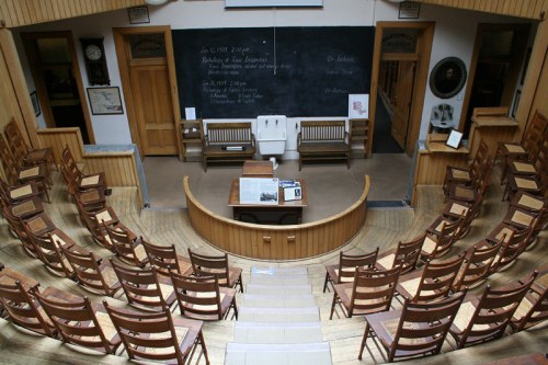 A historic classroom with wooden chairs arranged in a semicircle around a central desk and chalkboard.