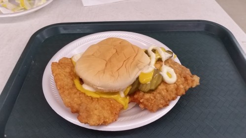 A plate with a sandwich featuring a bun, fried fish fillet, pickles, and condiments on a green tray.