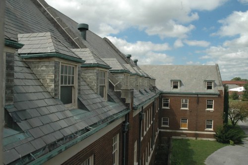 View of buildings with slate roofs and brick walls under a cloudy sky, featuring a grassy area in the foreground.