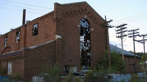 Abandoned brick building with broken windows, surrounded by overgrown vegetation and power lines.