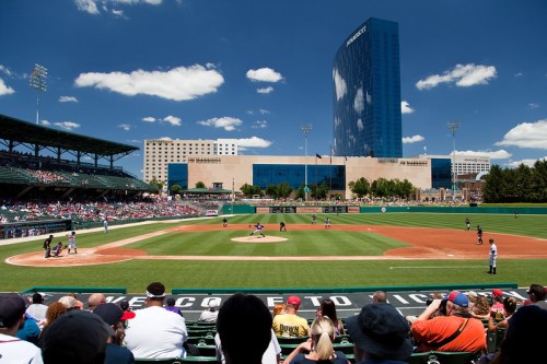 A sunny baseball stadium filled with fans, featuring a game in progress and a modern building in the background.