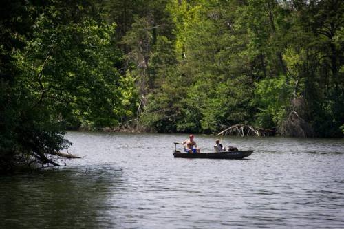Two people in a small boat fishing on a calm lake surrounded by lush green trees.