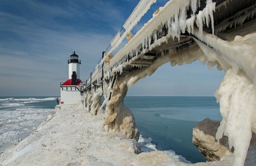 A frozen lighthouse with icicles, surrounded by snow and ice, against a blue sky and calm water.