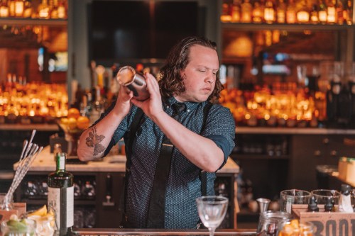 A bartender with curly hair shakes a cocktail shaker behind a bar filled with bottles and glasses.