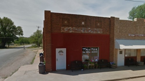 A red brick building with a sign reading "TWISTER The Movie MUSEUM" next to a tan building, on a quiet street.