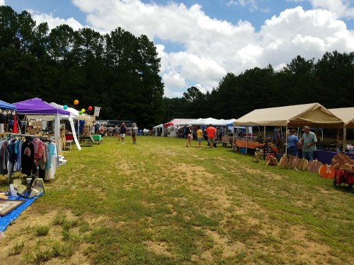 A vibrant outdoor market with colorful tents, vendors, and shoppers on a grassy path under a partly cloudy sky.