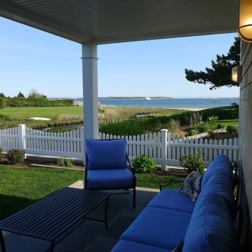 A serene outdoor patio with blue chairs, a table, and a view of the ocean and green landscape under a clear sky.