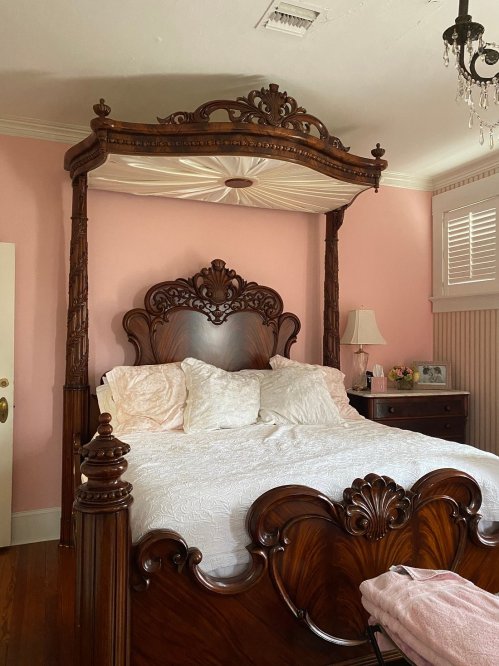 A beautifully crafted wooden four-poster bed with a white quilt in a softly lit pink bedroom.