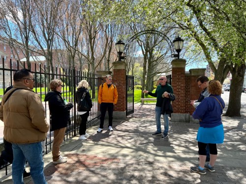 A group of people stands by a gated entrance, surrounded by trees and greenery, engaged in conversation on a sunny day.