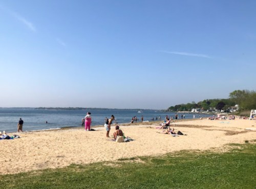 A sunny beach scene with people relaxing on the sand and swimming in the water, surrounded by greenery and blue skies.