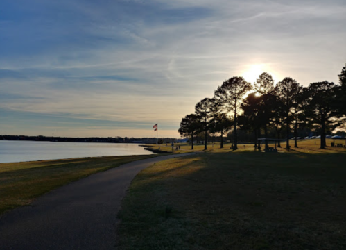 A serene lakeside scene at sunset, featuring a path, trees, and an American flag in the distance.
