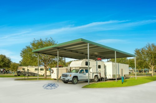 A covered parking area for RVs and a truck, surrounded by green grass and trees under a clear blue sky.