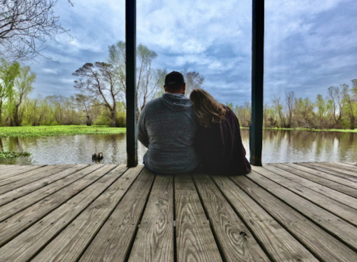 A couple sits together on a wooden dock, overlooking a serene lake surrounded by trees under a cloudy sky.